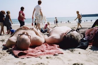 Two people are sunbaking at a beach, whilst others walk past them. 